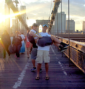 Jaxon Aaron, holding Brett Walton, pose on the Brooklyn Bridge with the evening sun setting behind the New York skyline.